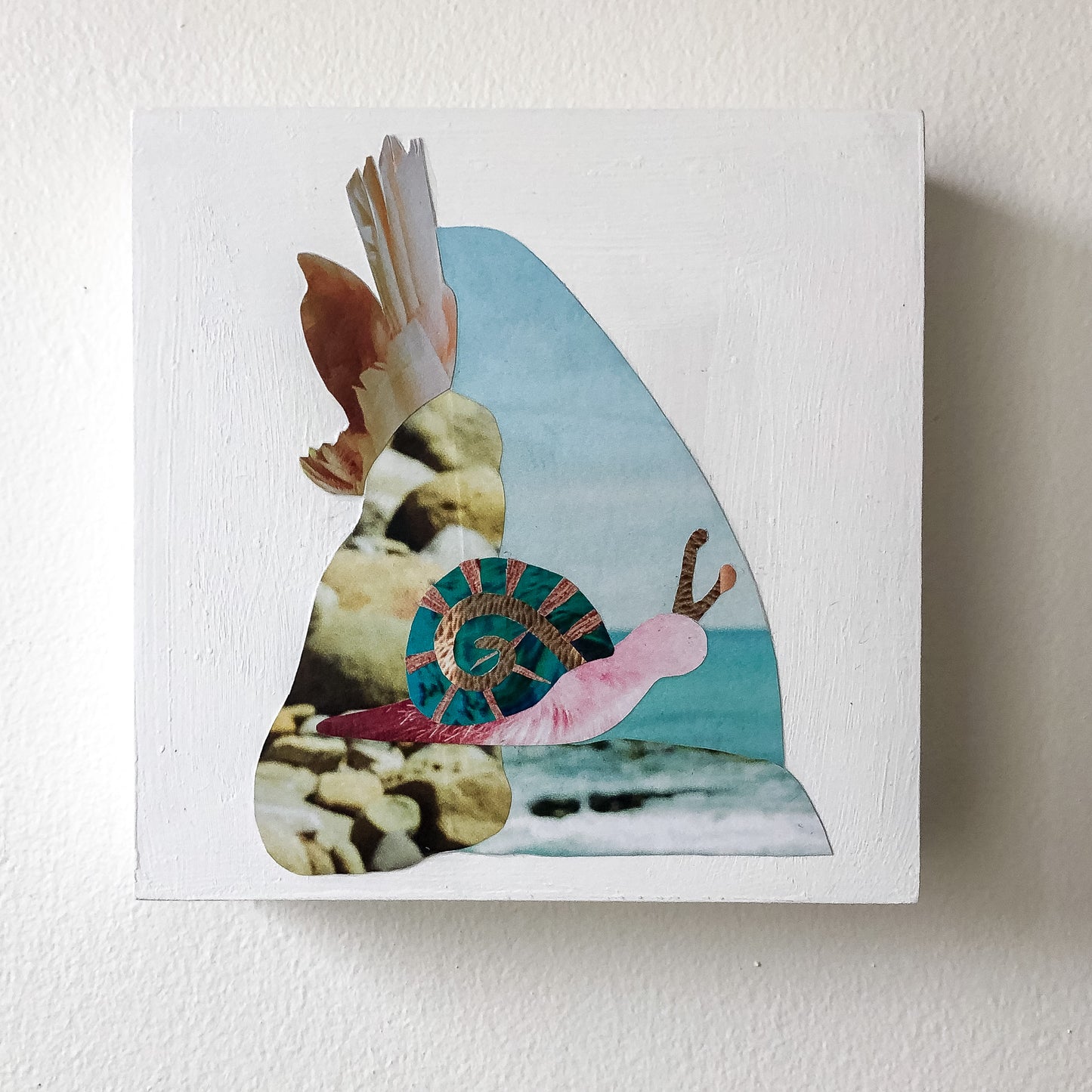 Analog collage art on a white painted square wood panel on a white wall.. Collage of a bech scene with a pink and blue textured snail.