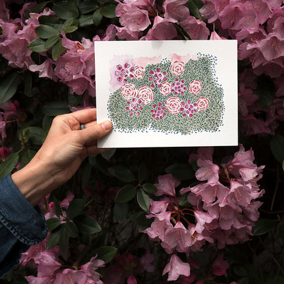 Hand holding floral watercolour painting in a garden in front of pink rhododendron flowers